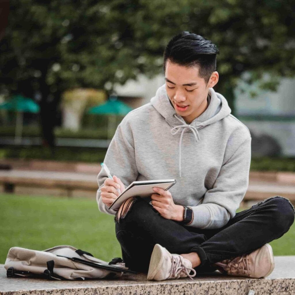 A teen sitting cross legged, looking down while writing on a notebook with a pen